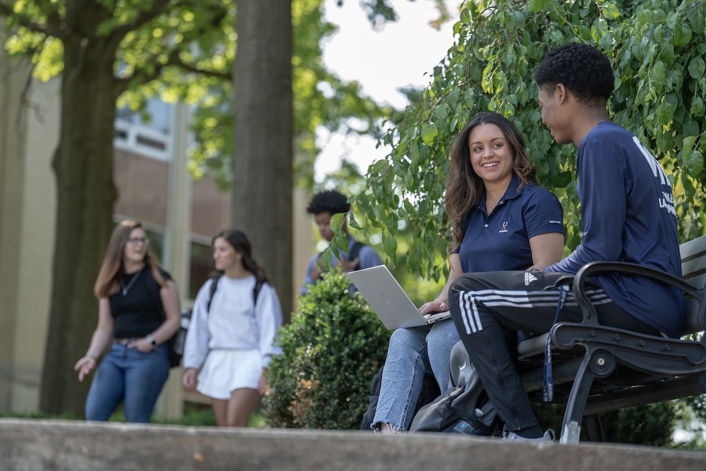 Two University of Akron students sit on an outdoor bench with a laptop, chatting, while other students walk by in the background on a sunny day.