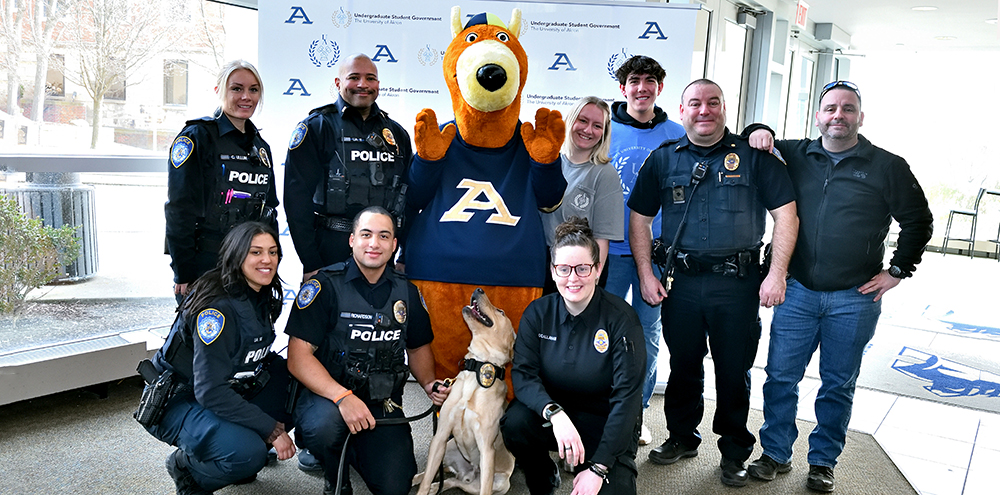 Gemini said A group of university police officers and students pose for a photo with Zippy, the University of Akron's kangaroo mascot, and a yellow lab police dog. The group is standing in front of a white backdrop featuring the University of Akron logo and the words 