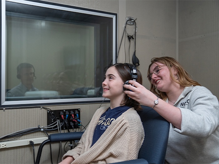 A woman helps a seated young woman put on headphones in a soundproof room, possibly for a hearing test. A man is visible through a window in the adjacent room.