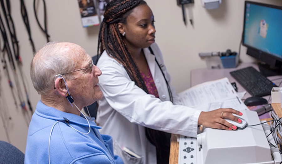 A healthcare professional operates audiology equipment while assisting an older man wearing hearing test headphones in a medical office.