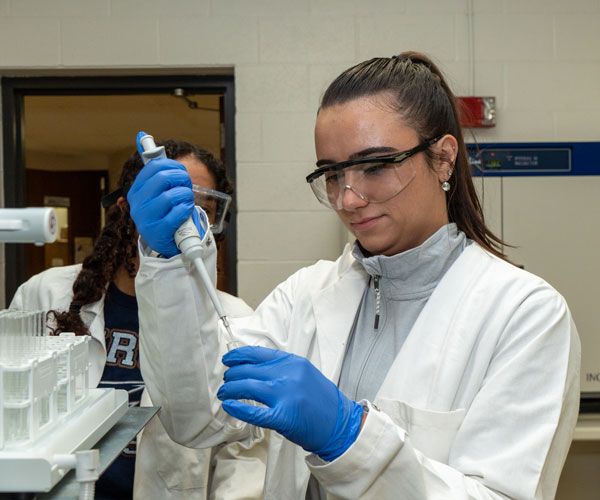 caucasian woman with long brown hair tied in ponytail wearing safety goggles using a pippet in a lab
