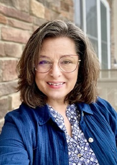 A woman with shoulder-length brown hair and glasses smiles at the camera. She is wearing a blue patterned blouse and a blue jacket, standing in front of a brick wall and window.