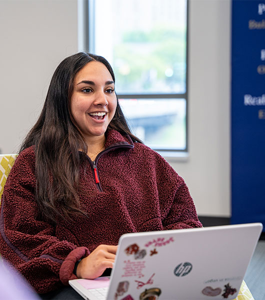 Student smiles while working on a laptop in a campus setting.