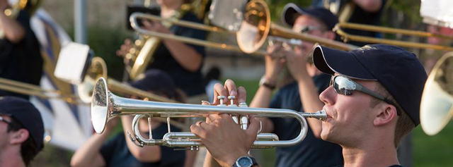 A group of musicians wearing sunglasses and hats play brass instruments, including trumpets and trombones, outdoors in bright sunlight.