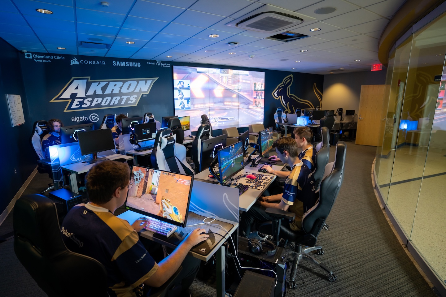 Several people wearing matching jerseys play video games on computers in a modern esports arena, with large screens showing game footage and Akron Esports logos on the walls.