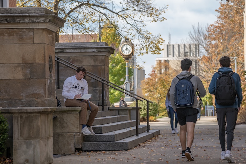 A student sits on stone steps studying, while two others with backpacks walk along a tree-lined campus path on a fall day. A campus clock and autumn foliage are visible in the background.
