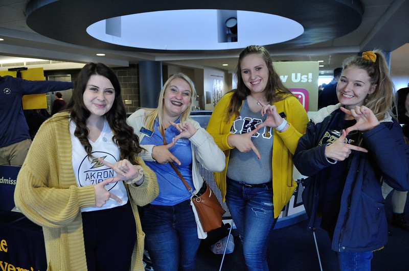 Four female students stand together indoors, smiling and posing for a photo. Each student is using their hands to form a stylized 