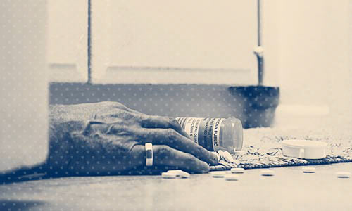 A hand on a floor with a pill bottle nearby and pills on the floor