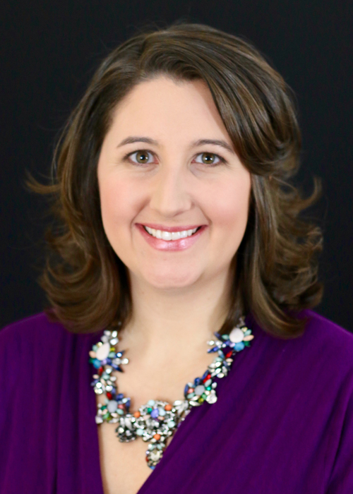 Photo of woman in purple top against and smiling against dark background.